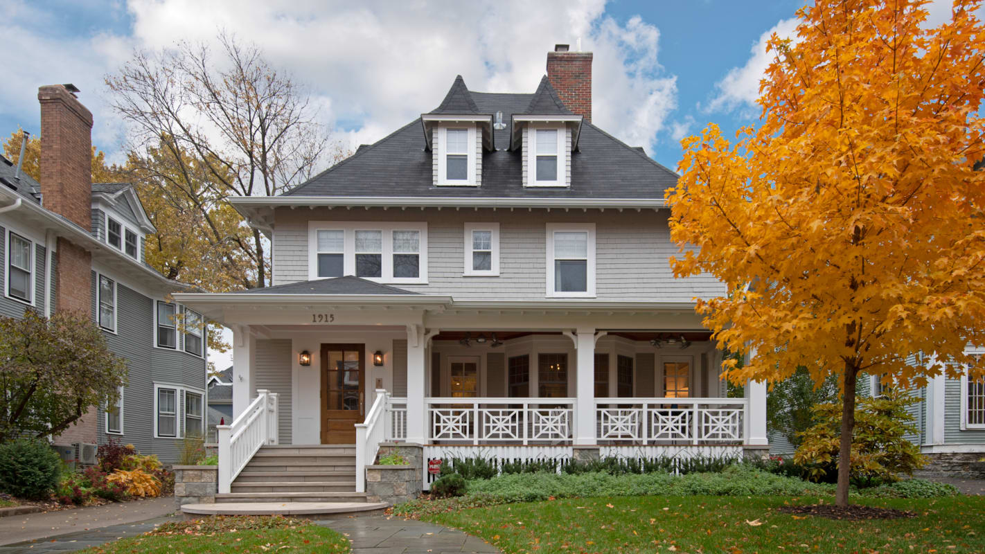 Charming two-story home with wraparound porch nestled in quiet residential neighborhood, surrounded by vibrant autumn foliage and mature trees.