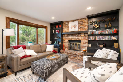 Interior of a cozy living room featuring a leather sofa, a patterned ottoman, and a brick fireplace.
