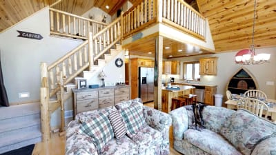 Interior of a rustic cabin with wooden stairs, plush sofas, and a kitchen area.
