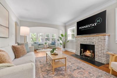 Cozy living room with a brick fireplace, a television, and natural light from bay windows.