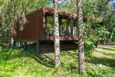 A contemporary brown cabin elevated on stilts in a wooded area with a deck and chairs.