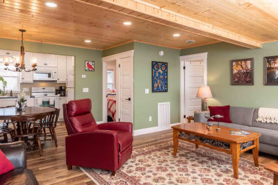 A cozy living room featuring a gray sofa, red recliner, and wooden coffee table on patterned rug, with a warm wood ceiling and a glimpse of the kitchen area.