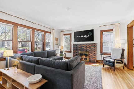 A bright living room with a sectional sofa, large windows, a brick fireplace, and a mounted television.