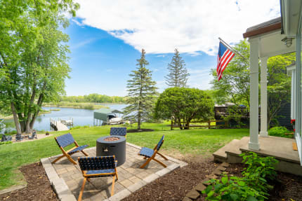 Outdoor seating area with a fire pit overlooking a lake, surrounded by trees and an American flag.