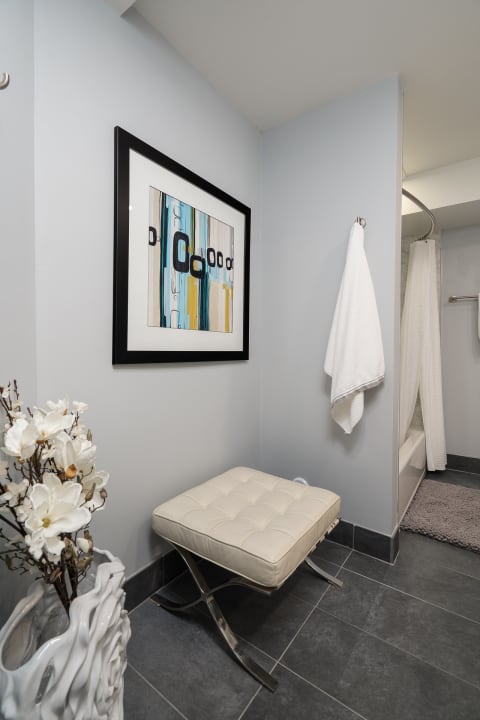 Modern bathroom corner featuring a tufted ottoman, framed artwork, and white flowers.