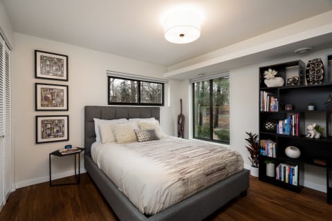 A modern bedroom featuring a stylish bed, artwork, and a black bookshelf, illuminated by natural light.