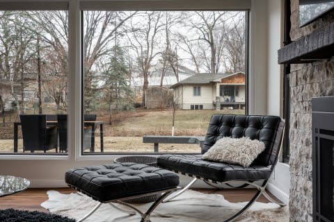 A stylish living room featuring a black lounge chair, ottoman, and a view of an outdoor area with trees and a house.