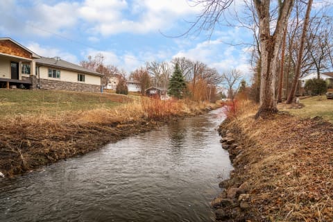 A creek winding between residential houses with dry grass and trees on either side.