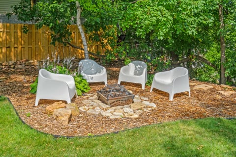 An outdoor seating area featuring white chairs and a stone fire pit amid lush greenery.