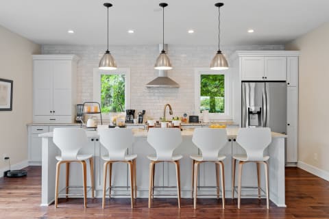 Modern kitchen with white bar stools, pendant lights, and a view of greenery.