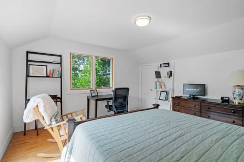 A bright bedroom with a bed, desk, rocking chair, and greenery outside the window.