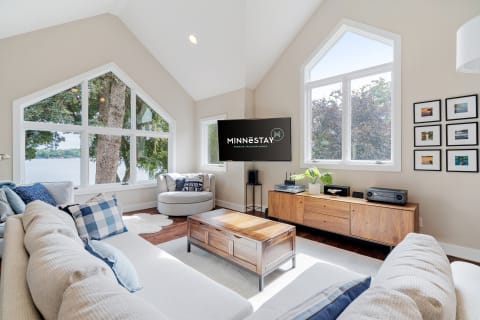 A modern living room with large windows overlooking a lake, featuring a white sofa, gray armchair, and wooden furniture.