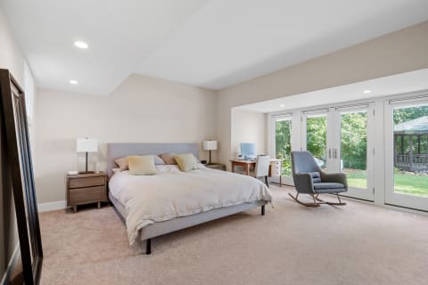 Minimalist bedroom with a bed, nightstand, desk, and a rocking chair next to large windows.