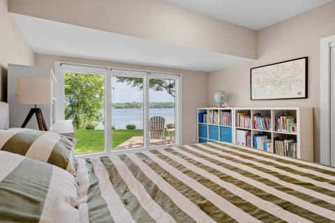 A cozy bedroom featuring a striped bedspread, large sliding doors to a lake view, and a colorful bookshelf.