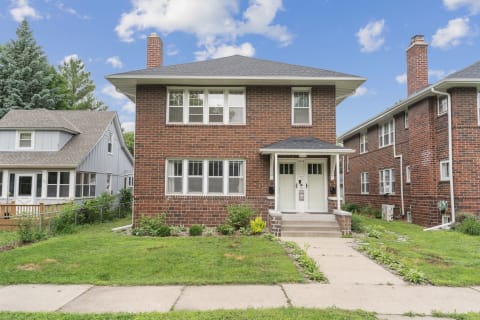 A brick duplex featuring two doors and green grass in front, with a gray house nearby.