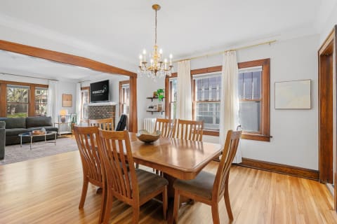 A dining area featuring a wooden table and chairs with a chandelier overhead, leading to a living room with a grey sofa and a brick fireplace.