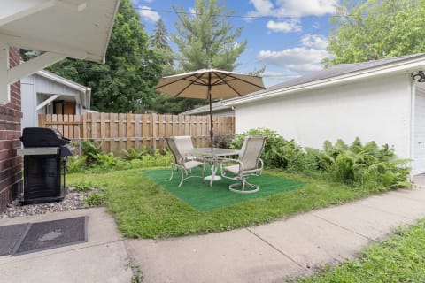 Outdoor patio area with a table and chairs under an umbrella, featuring a grill and lush greenery.