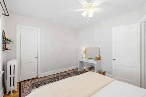 A well-lit bedroom featuring a white bed, a desk with a gold-framed mirror, and a patterned rug.