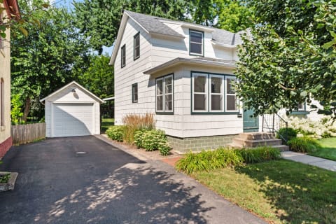 A two-story white house with green trim, a bay window, and a detached garage in a sunny environment.