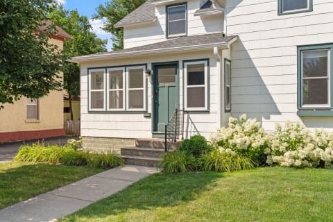 Front view of a two-story house with white siding and green accents, featuring a porch and blooming hydrangeas.