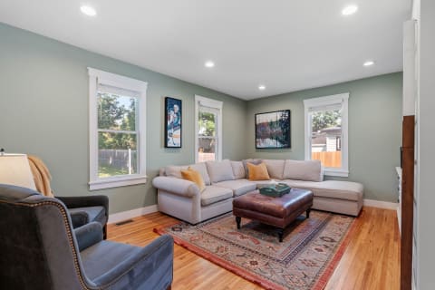 A modern living room featuring a gray sectional sofa, a plush brown ottoman, and sage green walls, illuminated by natural light.