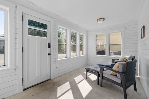 A sunlit sunroom featuring white shiplap walls, wicker chairs, and large windows.