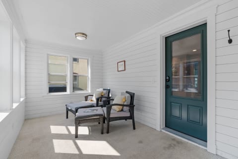 A bright sunroom with shiplap walls, two gray chairs, and a teal front door.