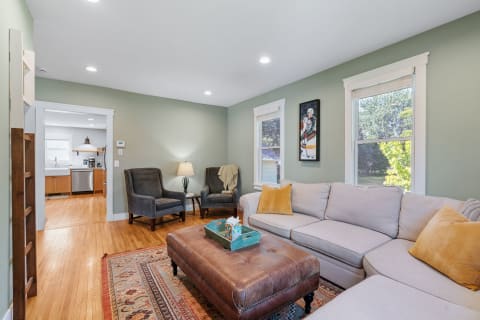 Living room with light green walls, gray sofa, leather ottoman, and natural light from windows.