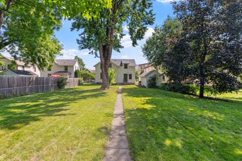 Backyard pathway leading to a two-story yellow house with trees and a lawn.