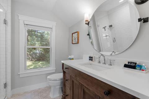 A sleek bathroom featuring a circular mirror, wooden vanity, and large window with greenery outside.