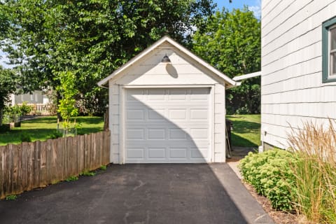 Small white garage with a closed door and lush greenery in the background.