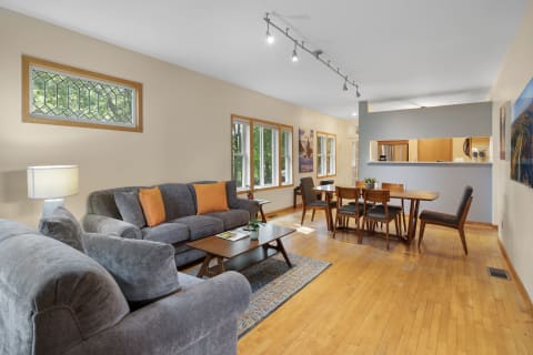 A spacious living room and dining area featuring gray sofas, orange cushions, wooden dining table, and large windows.