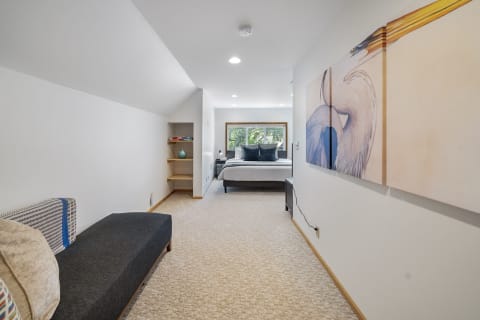 A minimalist bedroom with a gray chaise lounge, a bed with pillows, and a colorful bird artwork on the wall.