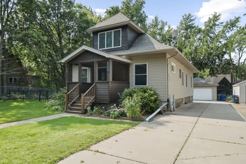 A two-story house with a brown exterior and a porch surrounded by greenery.