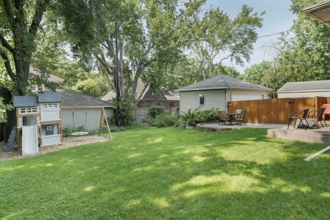 A green backyard with a playhouse and swing set, featuring a seating area and trees.