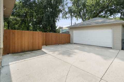 Outdoor area with a driveway, garage, and wooden fence surrounded by trees.