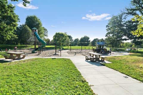 Playground with a green slide, swings, and a playhouse surrounded by grass and trees.