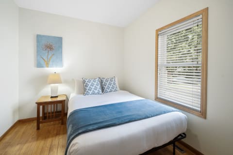 A cozy bedroom featuring a double bed, side table with lamp, and a window with a garden view.