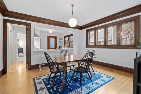 A dining area with a wooden table, black chairs, and large windows, featuring warm lighting and a colorful rug.