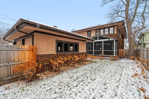 A winter view of a home with snow, featuring beige walls and brick details, surrounded by a wooden fence.