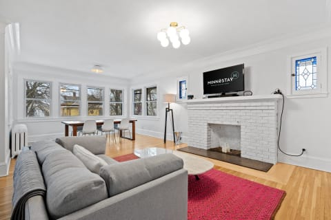 A modern living room featuring a gray sofa, wood dining table, white brick fireplace, and large windows.