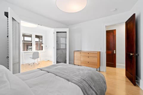 A modern bedroom with a bed, dresser, and a glimpse of a workspace through glass doors.