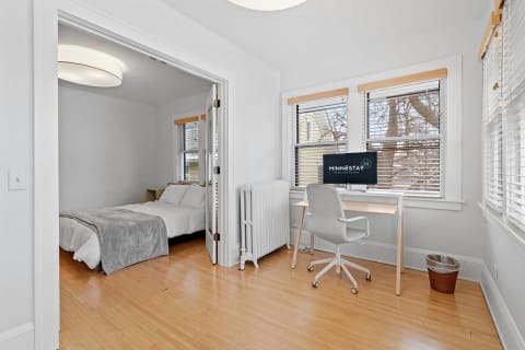 A room featuring a bed, desk, and chair with large windows and wooden flooring.