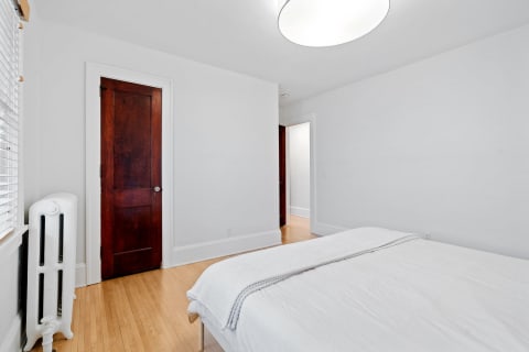 A serene bedroom featuring a made bed, wooden door, and radiator, with natural light filtering through the window.