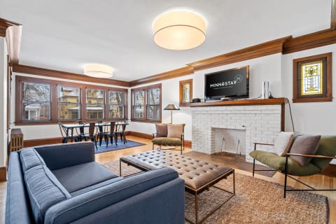 A modern living room featuring a blue couch, tufted leather ottoman, and a dining area with black chairs.