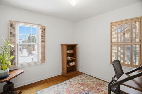 A cozy reading nook featuring a window with shutters, a wooden bookshelf, and a round table with a potted plant.