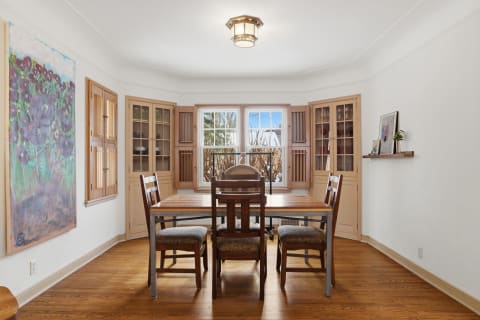Dining area with a wooden table, upholstered chairs, vibrant painting, and natural light from windows.