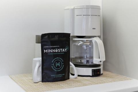 A modern coffee setup including a white coffee maker, a black coffee bag from MINNESTAY, and a white ceramic mug on a woven mat.