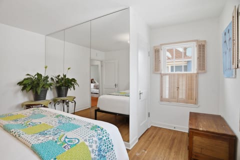 A bright bedroom with a bed adorned in a colorful quilt, mirrored wardrobes, and a shuttered window.