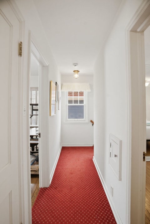 A hallway featuring white walls and a red carpet, illuminated by natural light and a vintage light fixture.